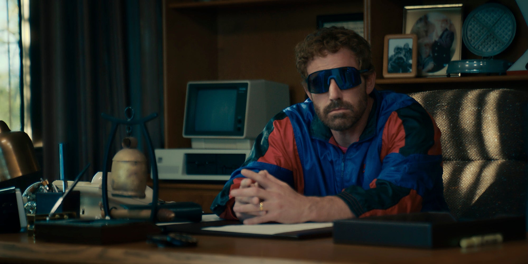 Ben Affleck wearing large sunglasses and a tracksuit sitting at a desk in the new movie AIR.