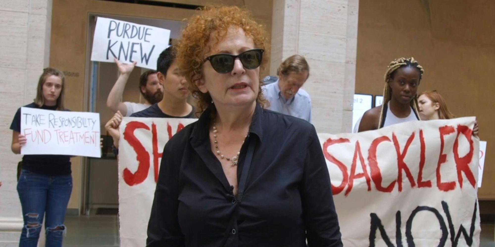 Nan Goldin speaking as people hold signs behind in All the Beauty and the Bloodshed