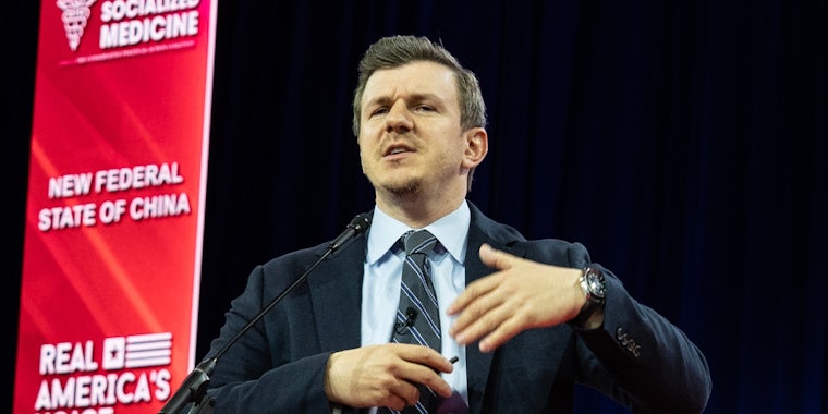 James O'Keefe speaking into microphone in front of dark blue and red background