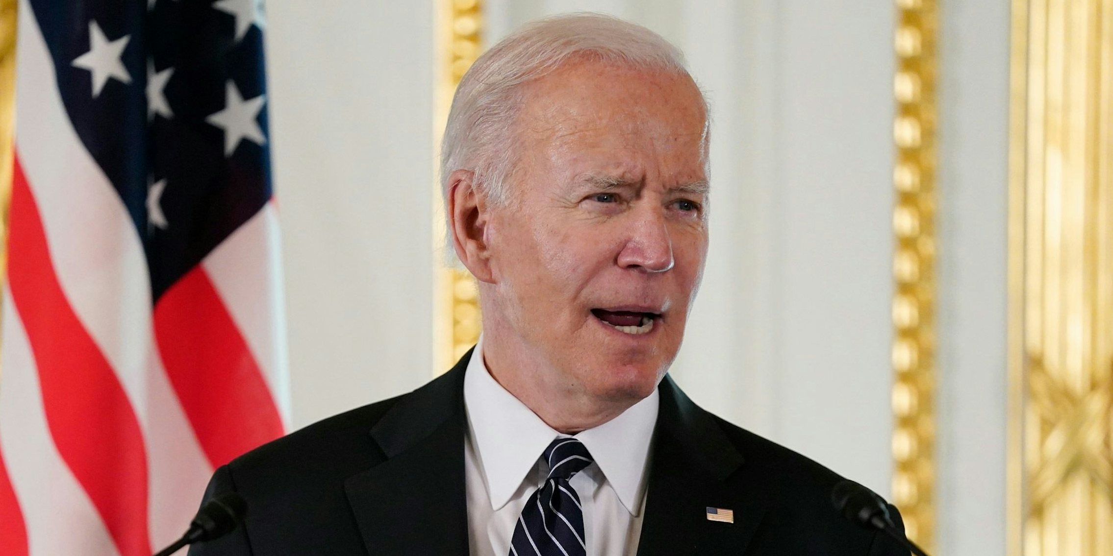 Joe Biden speaking into microphones in front of white and gold background with American flag to his left