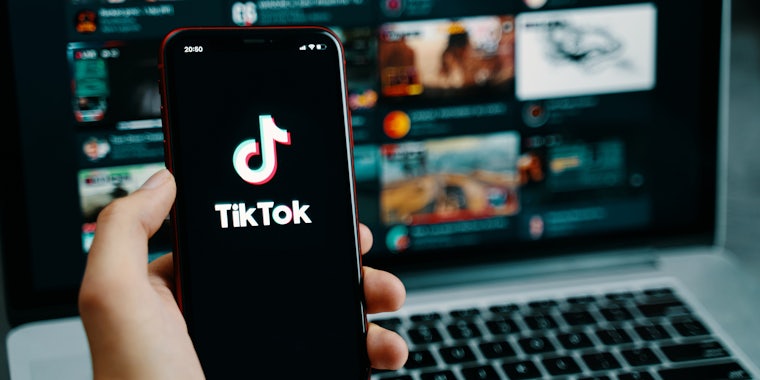hand holding phone with TikTok on screen in front of laptop