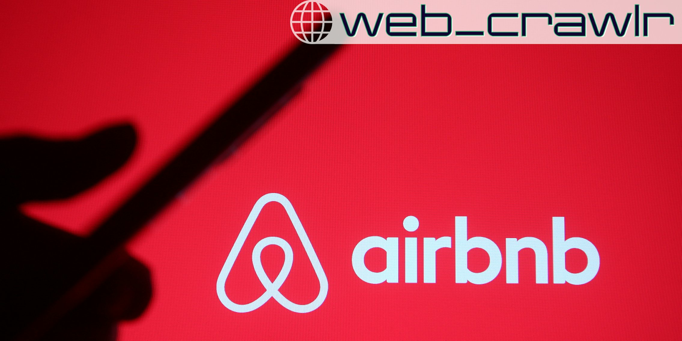 In this photo illustration Airbnb, Inc. logo is seen in front of a silhouette of a hand holding a mobile phone. The Daily Dot newsletter web_crawlr logo is in the top right corner.