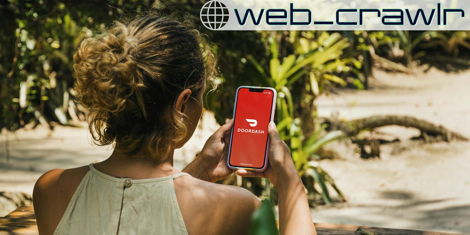 A woman holding a smartphone with the DoorDash logo on it. The Daily Dot newsletter web_crawlr logo is in the top right corner.