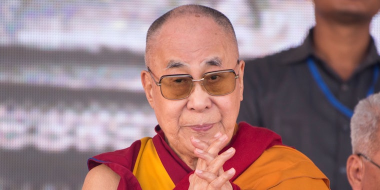 Tibetan spiritual leader the Dalai Lama with hands together in front of grey background