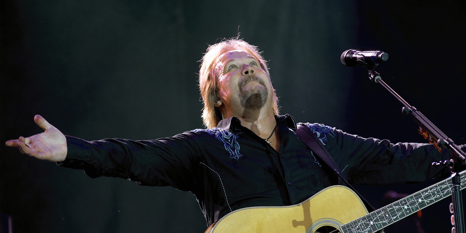 Travis Tritt with arms out with guitar and microphone in front of dark background