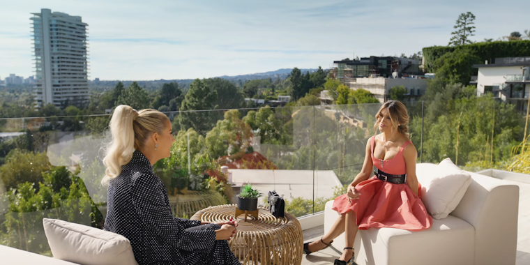 chrishell stause in a pink dress and belt, sitting on a couch opposite Emma Hernan in a embellished dark suit, on the rooftop of a house once owned by Harry Styles in Los Angeles