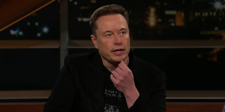 Elon Musk speaking with hand on chin in front of city background