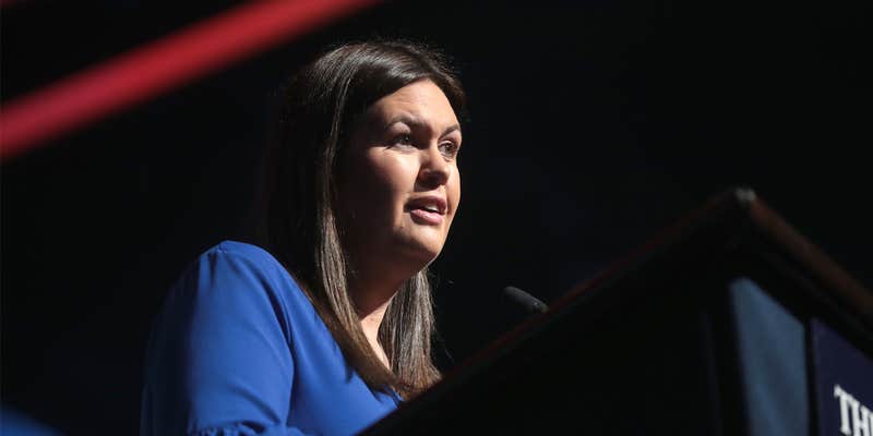 Secretary Sarah Huckabee Sanders speaking into microphone in front of dark background