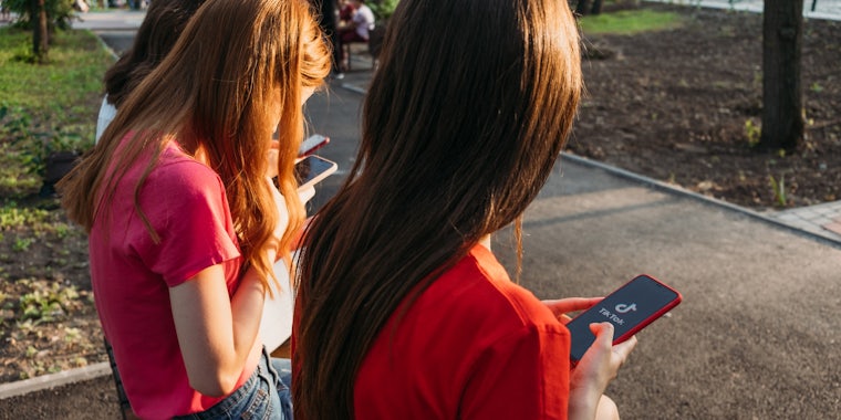 View from behind of two people with long hair, one brunette and one with red hair, sitting outside. They book look at the TikTok apps on their phones.