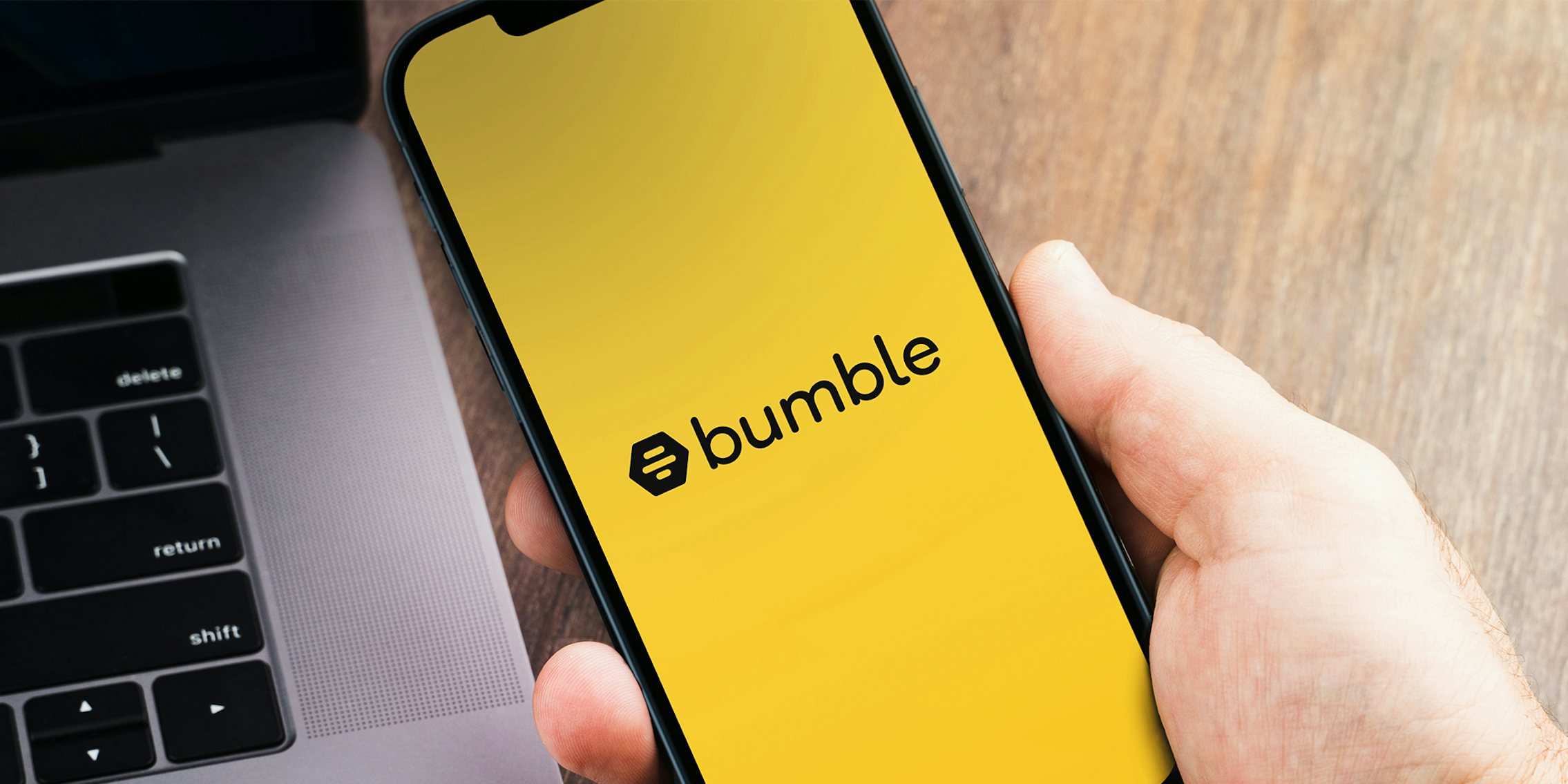 Male hand holding a smartphone with Bumble app on the screen with a computer beside it. Wooden Background.