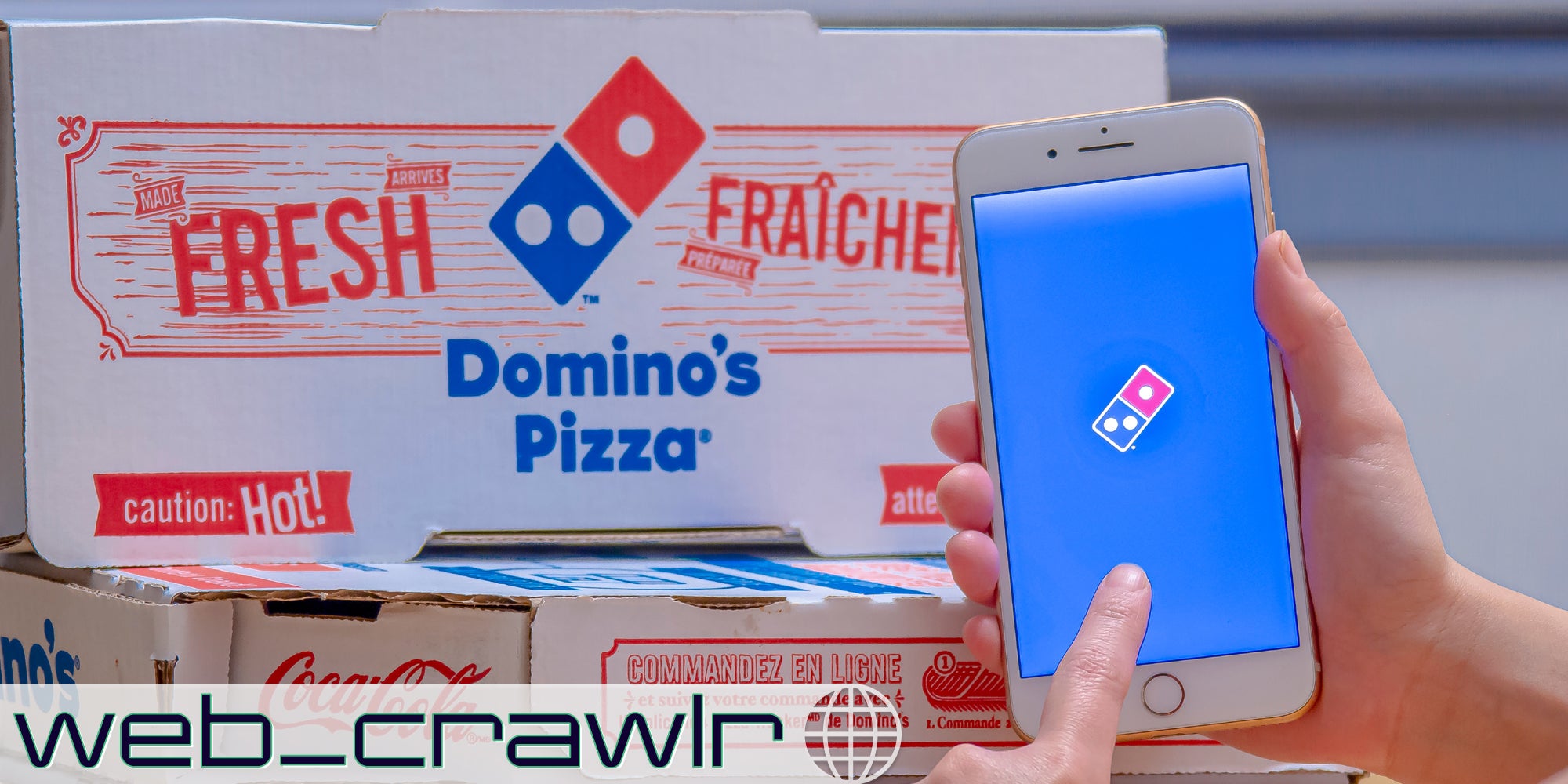 A phone with the Domino's app on it in front of boxes of Domino's Pizza. The Daily Dot newsletter, web_crawlr, logo is in the bottom left corner.