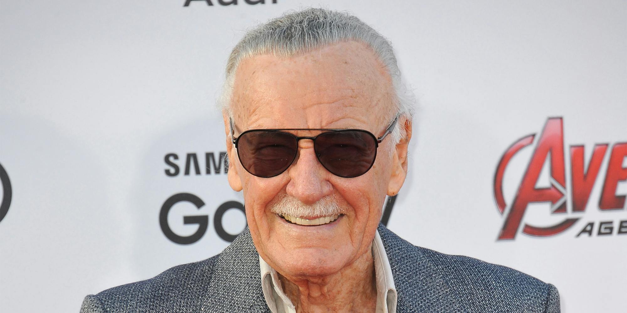 Stan Lee smiling in front of white Avengers background