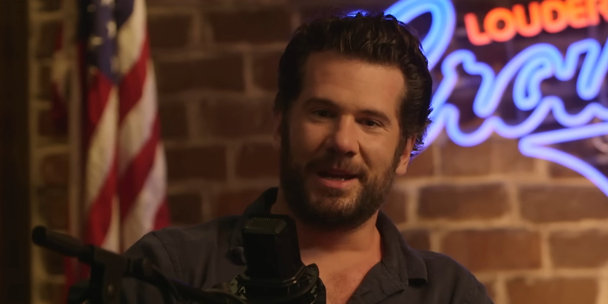 Steven Crowder speaking into microphone in front of brick background