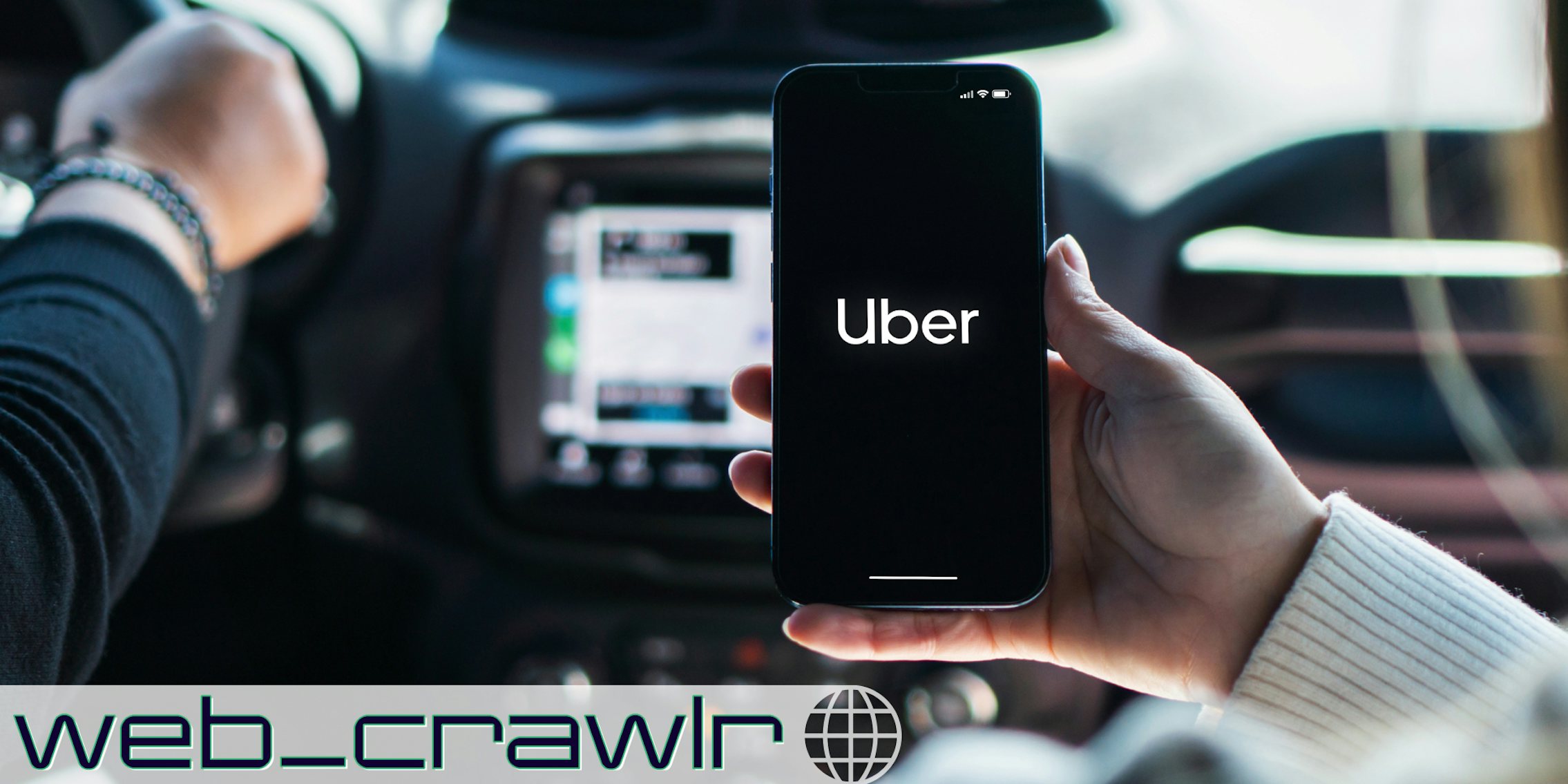 A person holding a phone with the Uber app on it while in a car. The Daily Dot newsletter web_crawlr logo is in the bottom left corner.