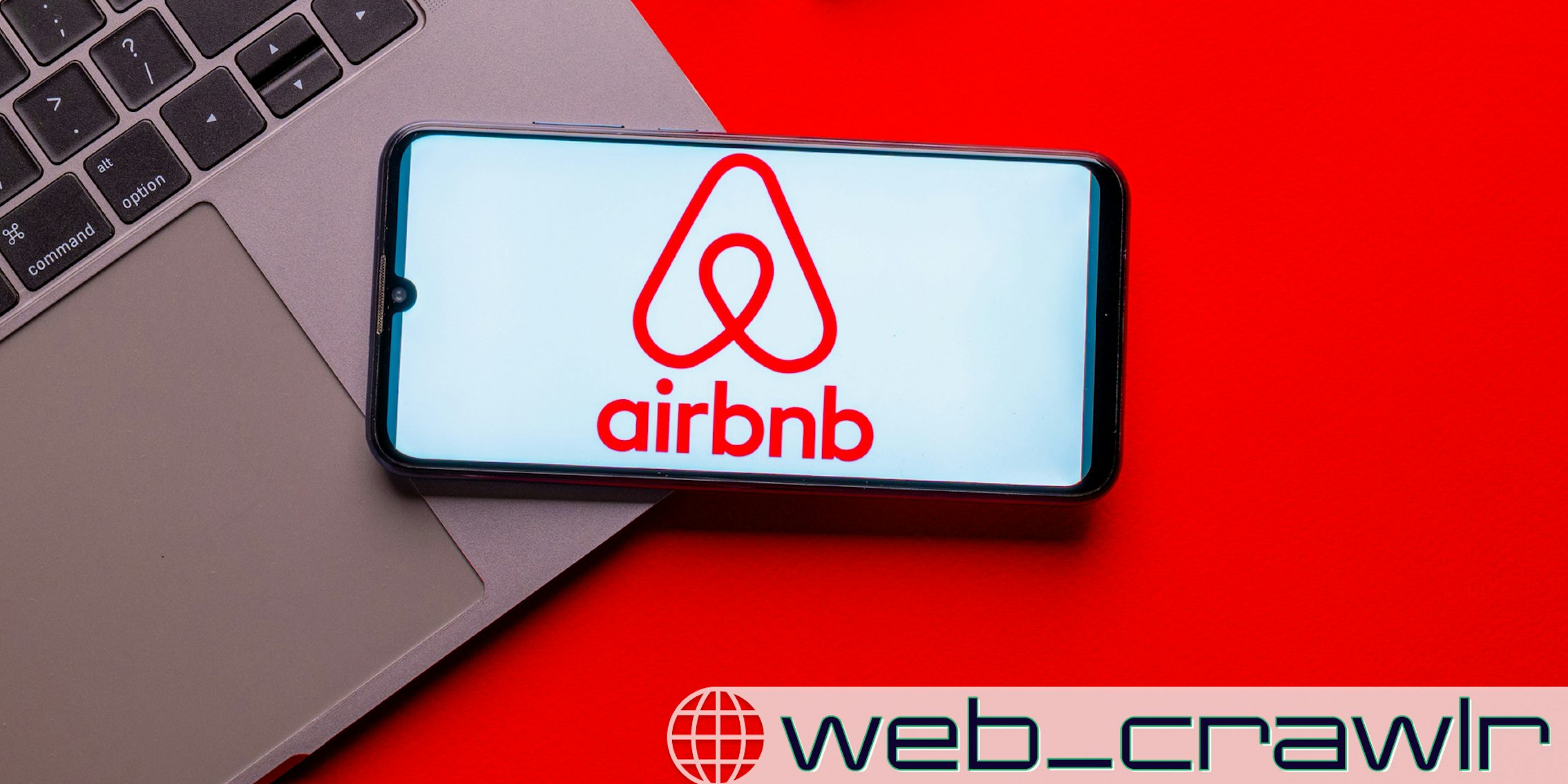 A phone with the Airbnb logo on it resting on a laptop. The Daily Dot newsletter web_crawlr logo is in the bottom right corner.