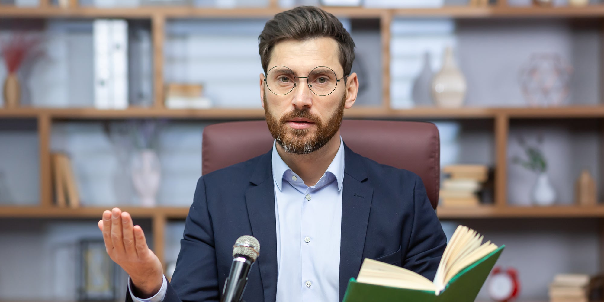Pastor Sitting and speaking into a microphone in front of the camera, holding a holy book.