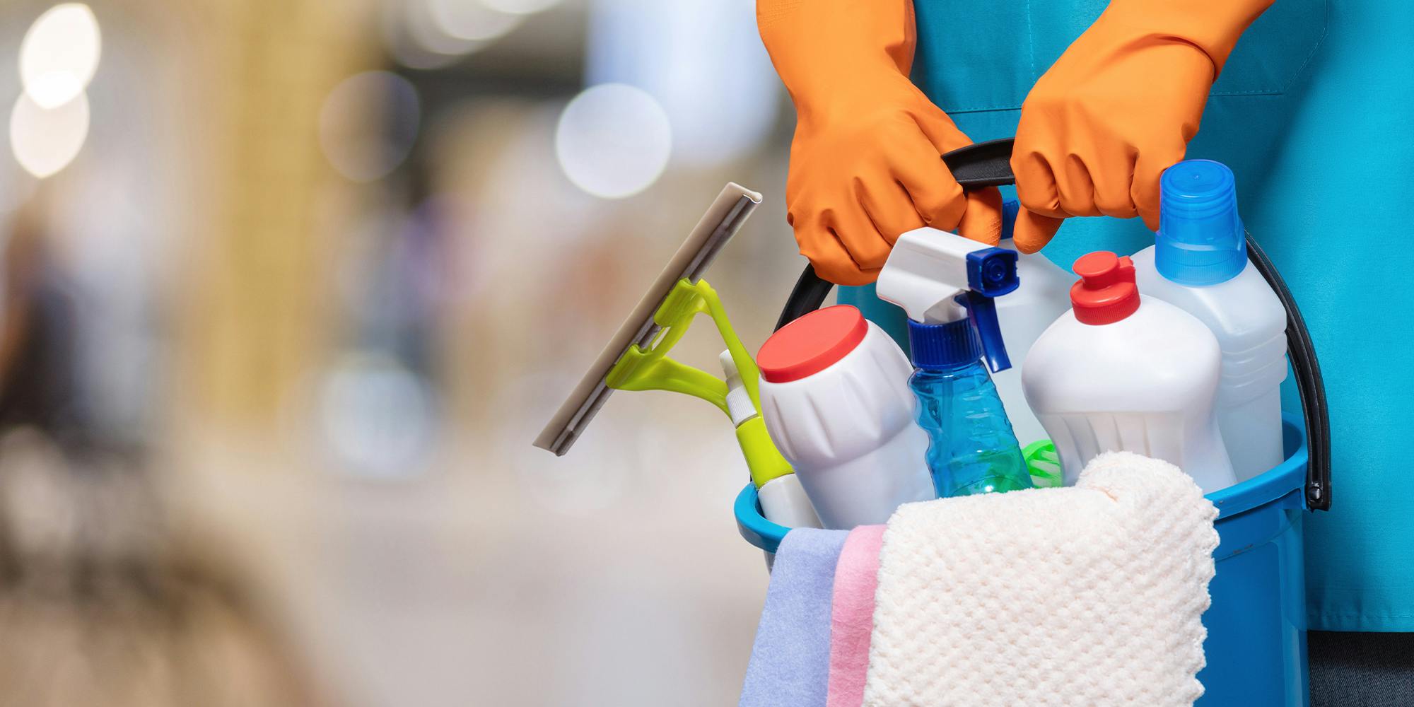 A bucket of cleaning products in hands with rubber gloves on a blurred background.