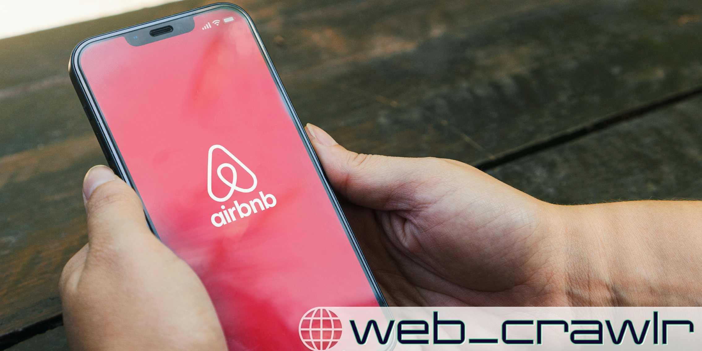 A person holding a phone with an Airbnb logo on it. The Daily Dot newsletter web_crawlr logo is in the bottom right corner.