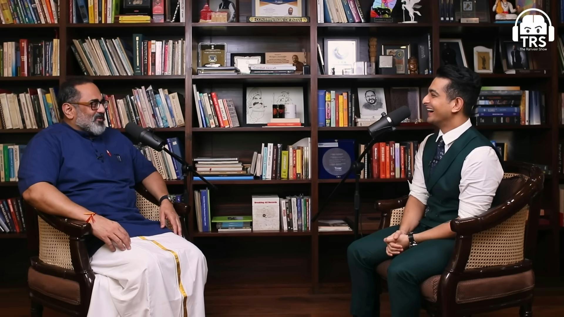 two men speaking in front of bookshelves