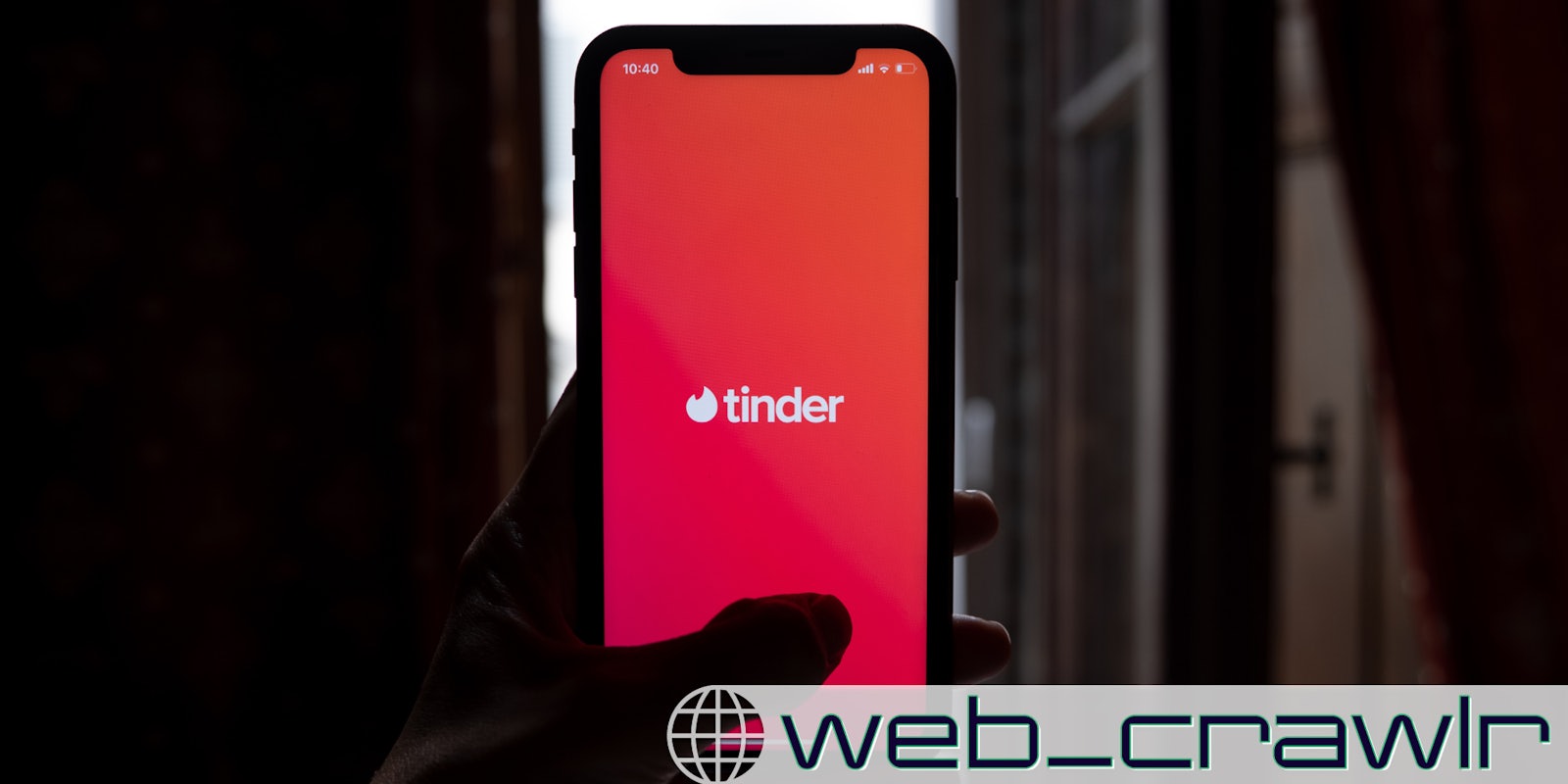 A person holding a smartphone with the Tinder logo on it. The Daily Dot newsletter web_crawlr logo is in the bottom right corner.