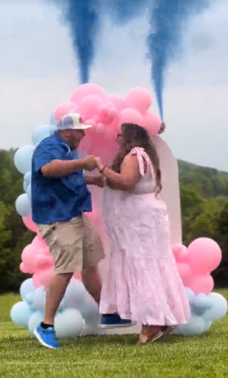 Excited couple Allie and Noah dressed in pink and blue jumping in excitement in front of pink and blue balloons as blue flares go off marking that they are going to have a boy.