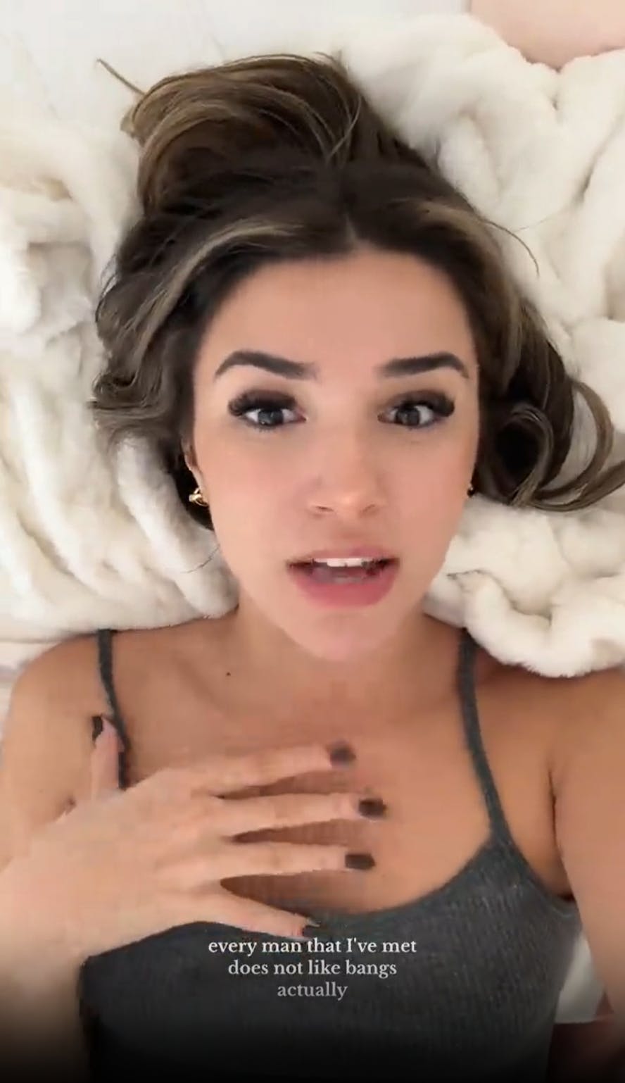 A brunette woman lying on a white fluffy blanket, holding her hand to her chest. Captions on the image read, 'every man that I've met does not like bangs actually.'