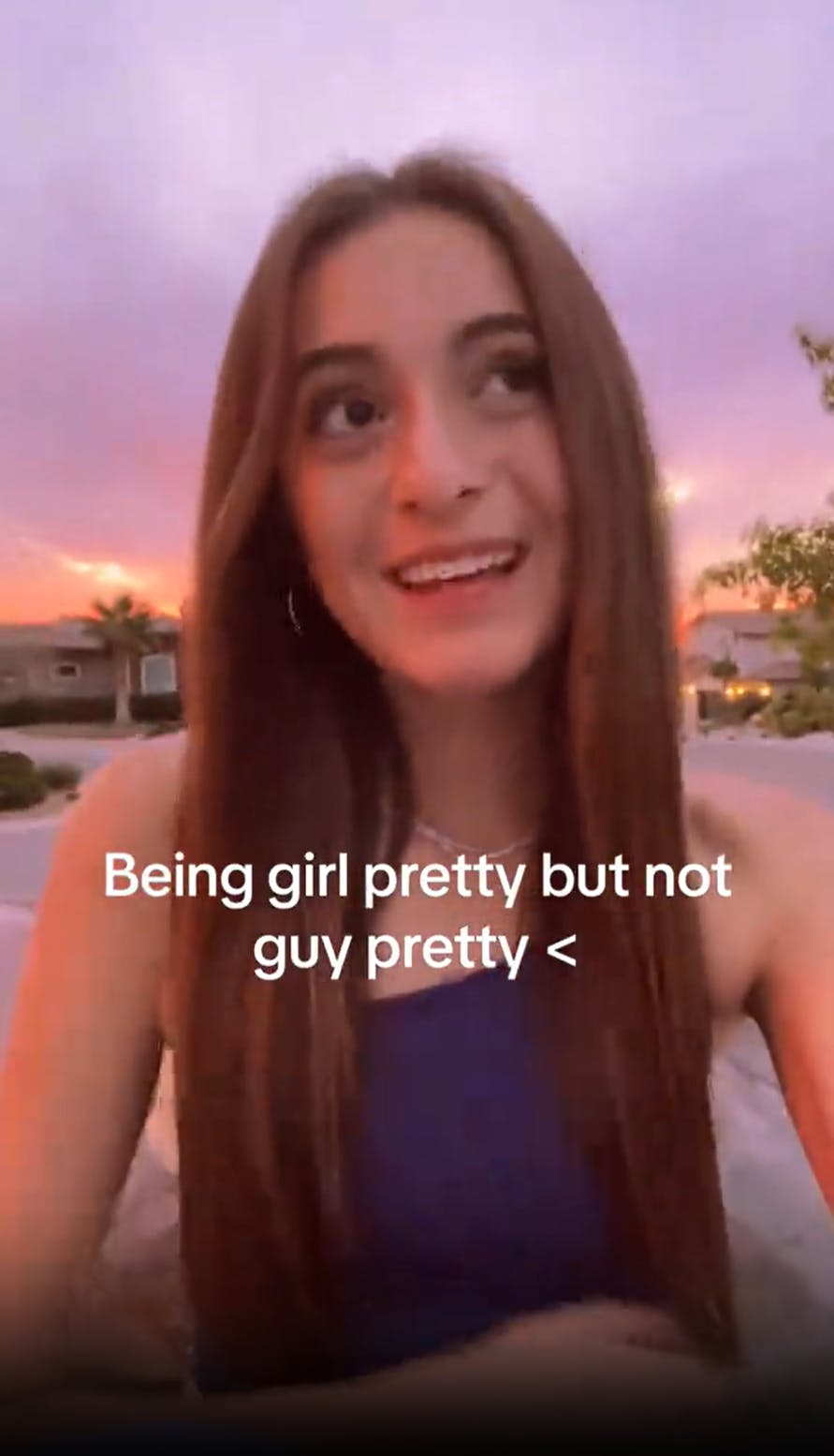 A white woman with long brown hair sitting on the ground in front of a sunset looking off to one side. Overlay text reads, 'Being girl pretty but not guy pretty