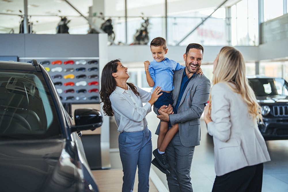 Happy family choosing new car, salesman showing them luxury auto at automobile dealership store. Customers selecting vehicle, consulting manager at modern showroom shop