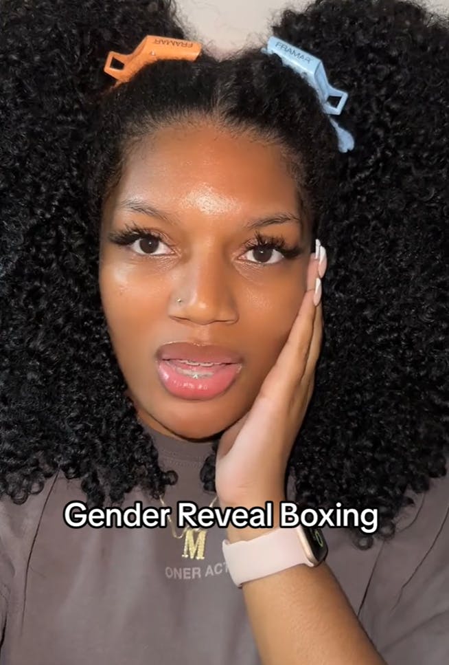 A black woman holding one hand to her face as she looks off-camera. Overlay text reads, 'Gender Reveal Boxing'