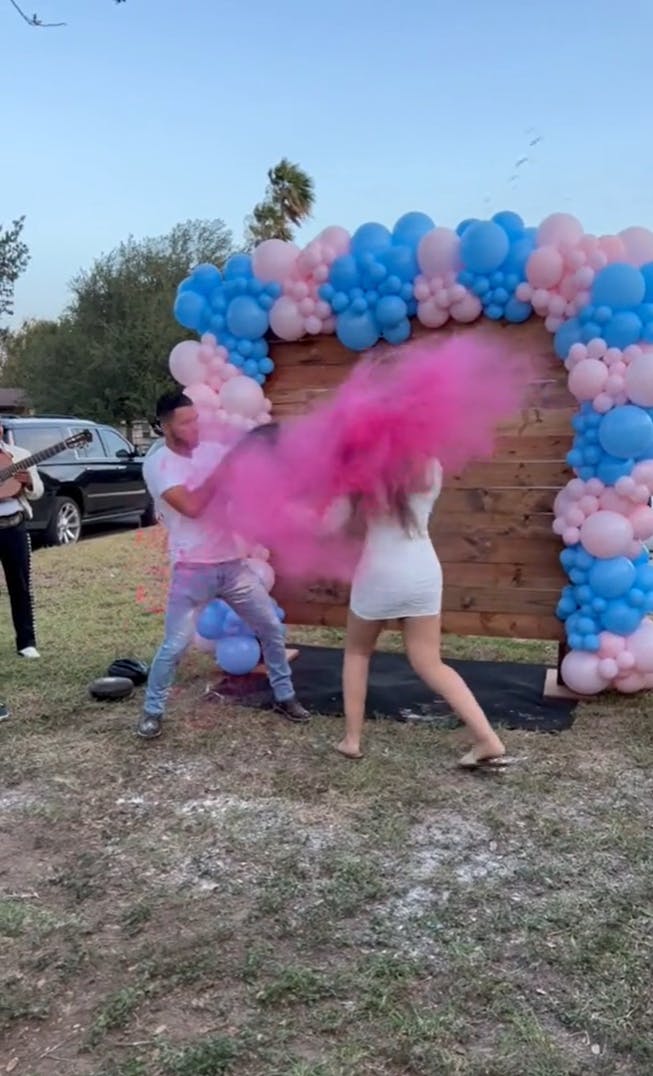 A man and woman surrounded by pink and blue balloons as the woman punches a mat and a poof of pink smoke blows into her face.