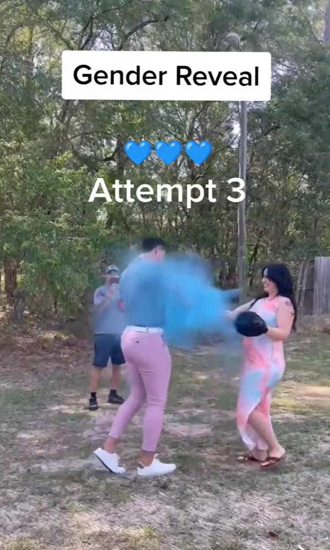 A couple in pink and blue clothes attempting a boxing punch gender reveal, a poof of blue smoke is shaken out of her strike mat. Text overlay reads, 'Gender Reveal Attempt 3' with three blue hearts.