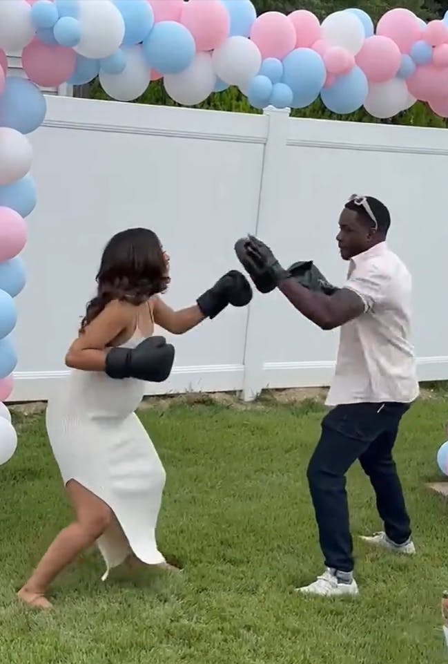 A man coaching his pregnant wife through how to punch strike mitts with boxing gloves to do a gender reveal. There is an archway of pink, blue, and white balloons around them.