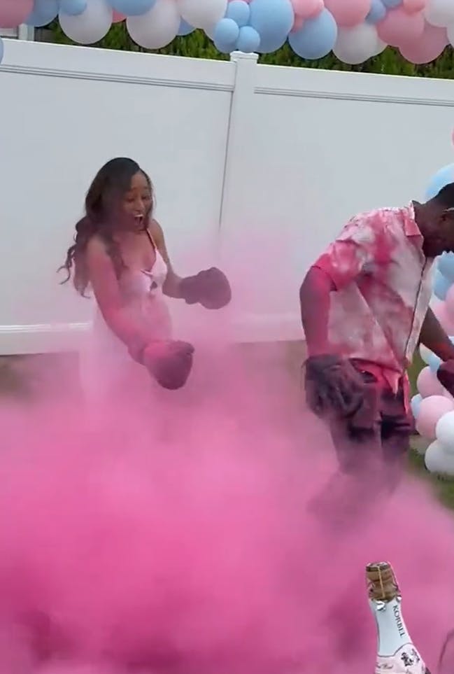 A couple in white clothing covered in pink smoke and dust after the pregnant partner punched a gender reveal bag on the man's strike mitt. She looks very excited.