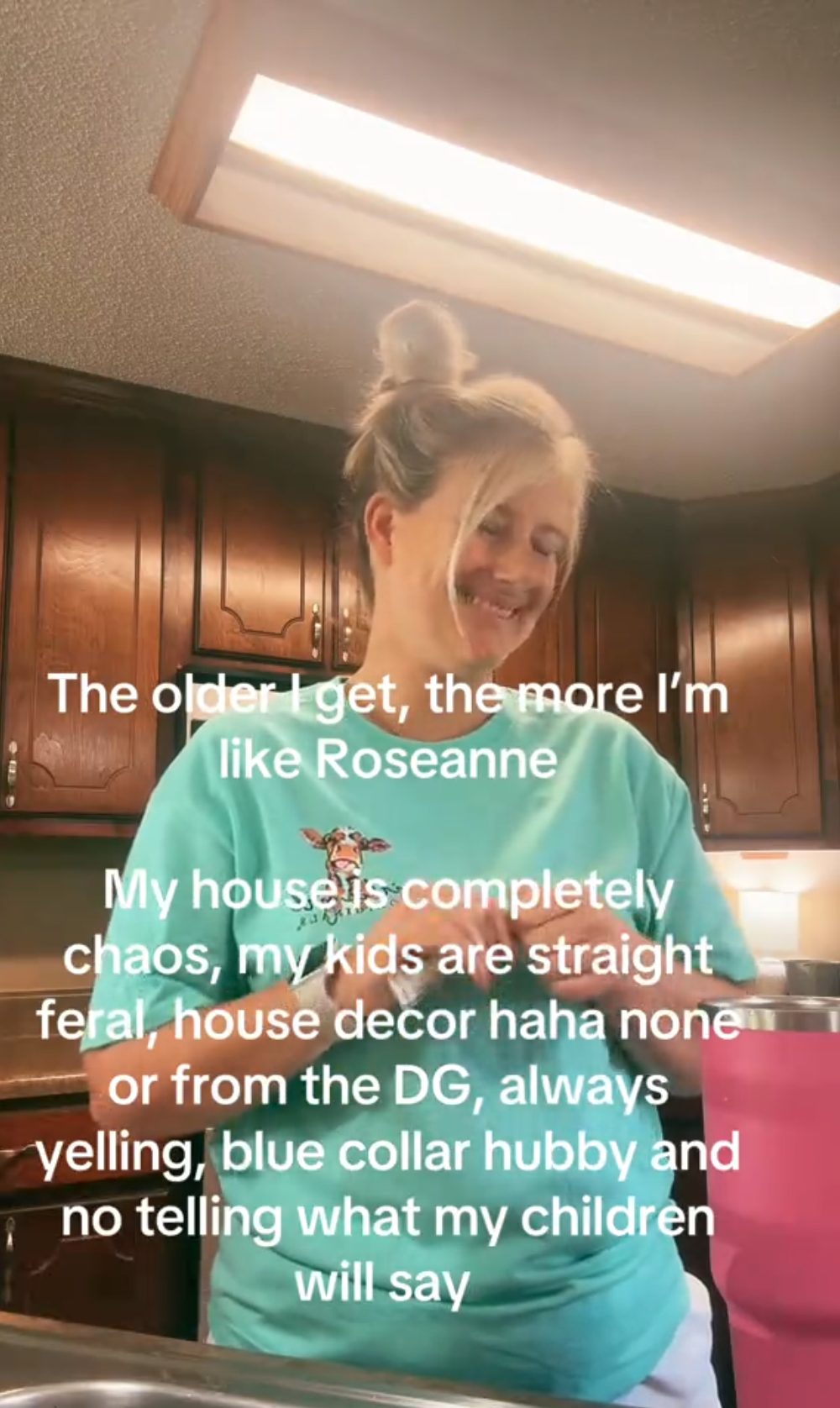 Screenshot of a blonde woman smiling in her kitchen as she prepares a drink. Text overlay reads, 'The older I get, the more I'm like Roseanne. My house is completely chaos, my kids are straight feral, house decor haha none or from the DG, always yelling, blue collar hubby and no telling what my children will say.'