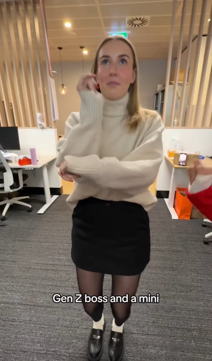 A Gen Z woman in a turtleneck and miniskirt dancing in the office with the text overlay, 'Gen Z boss and a mini'