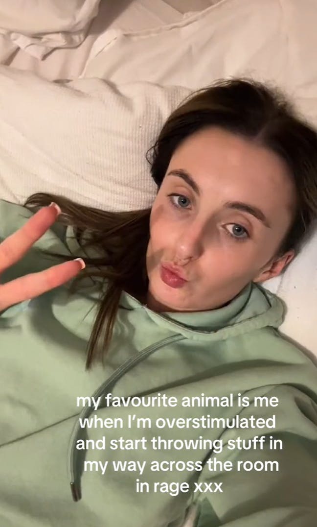 A brunette woman lying on a bed giving the camera a peace sign and duck face lips. Text overlay reads, 'My favourite animal is me when I’m overstimulated and start throwing stuff in my way across the room in rage xxx'