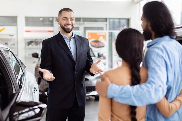 A cheerful car salesman is having a conversation with Indian couple inside a car dealership showroom