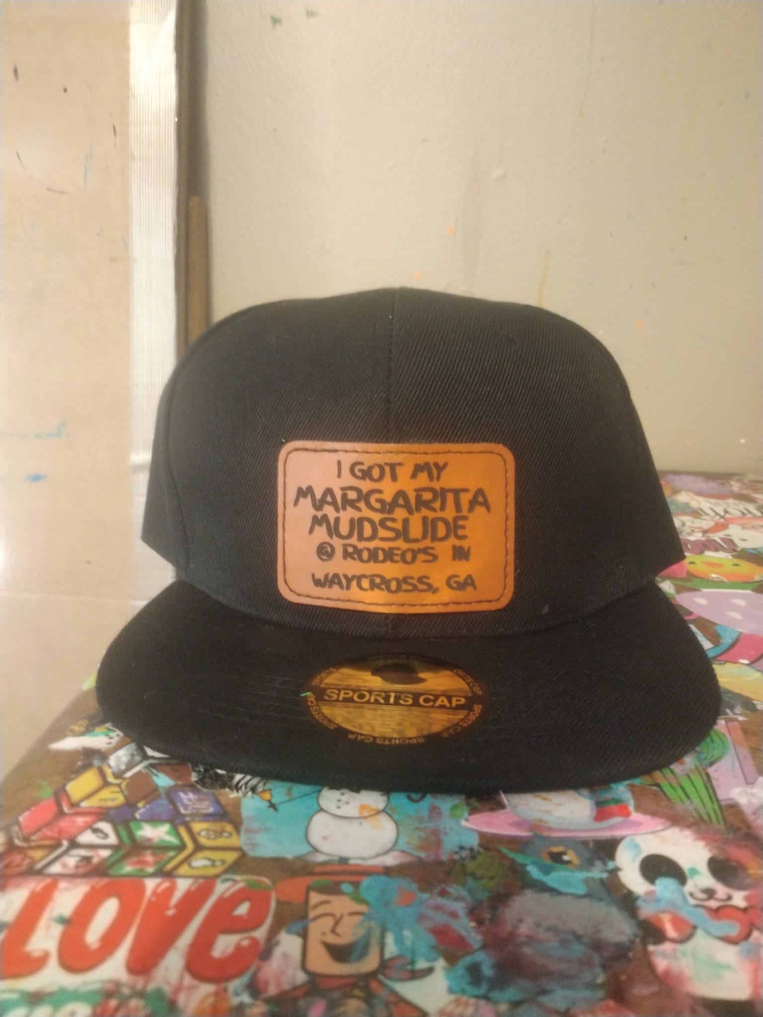 A black hat with a brown leather patch on it that reads, 'I got my margarita mudslide @ Rodeo's in Waycross, GA.'