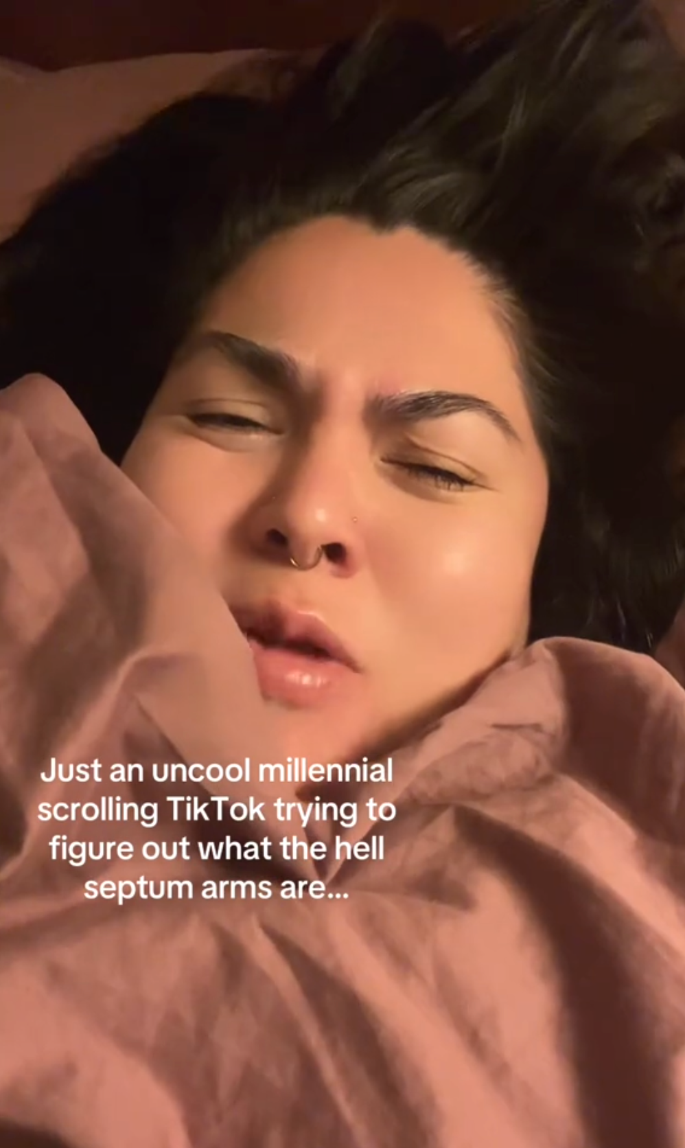 A woman cozy in a pink blanket looking confused. Text overlay reads, 'Just an uncool millennial scrolling TikTok trying to figure out what the hell septum arms are...'