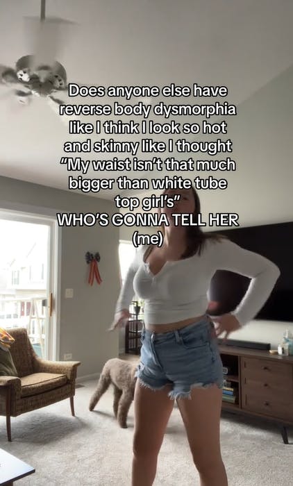 Video screenshot of a woman with text overlay saying, 'Does anyone else have reverse body dysmorphia like I think I look so hot and skinny like I thought 'My waist isn't that much bigger than white tube top girl's' WHO'S GONNA TELL HER (me)'