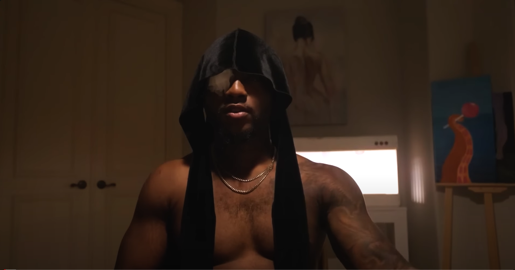 Screenshot of a video of a shirtless man wearing an untied durag on his head, a bandage covering one eye. There is dramatic overhead lighting.