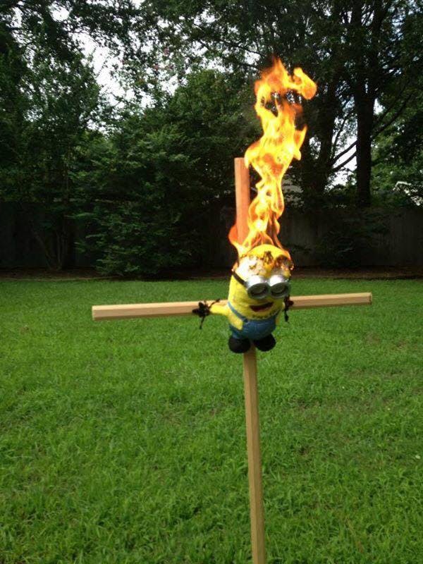 A Minion plush toy tied to a wooden cross in a yard, its head on fire.