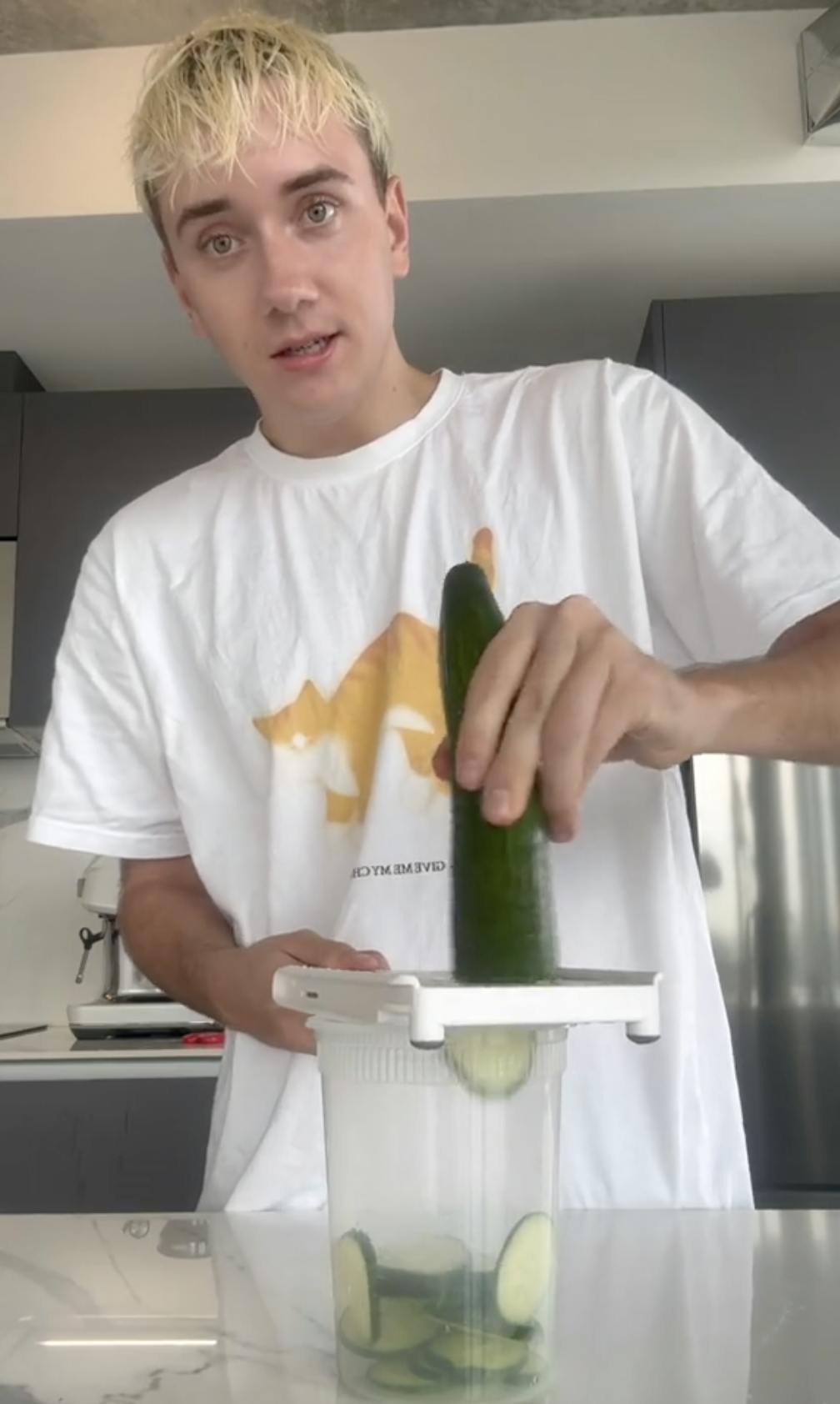 A young man with blond hair staring at the camera while slicing a cucumber for cucumber salad.