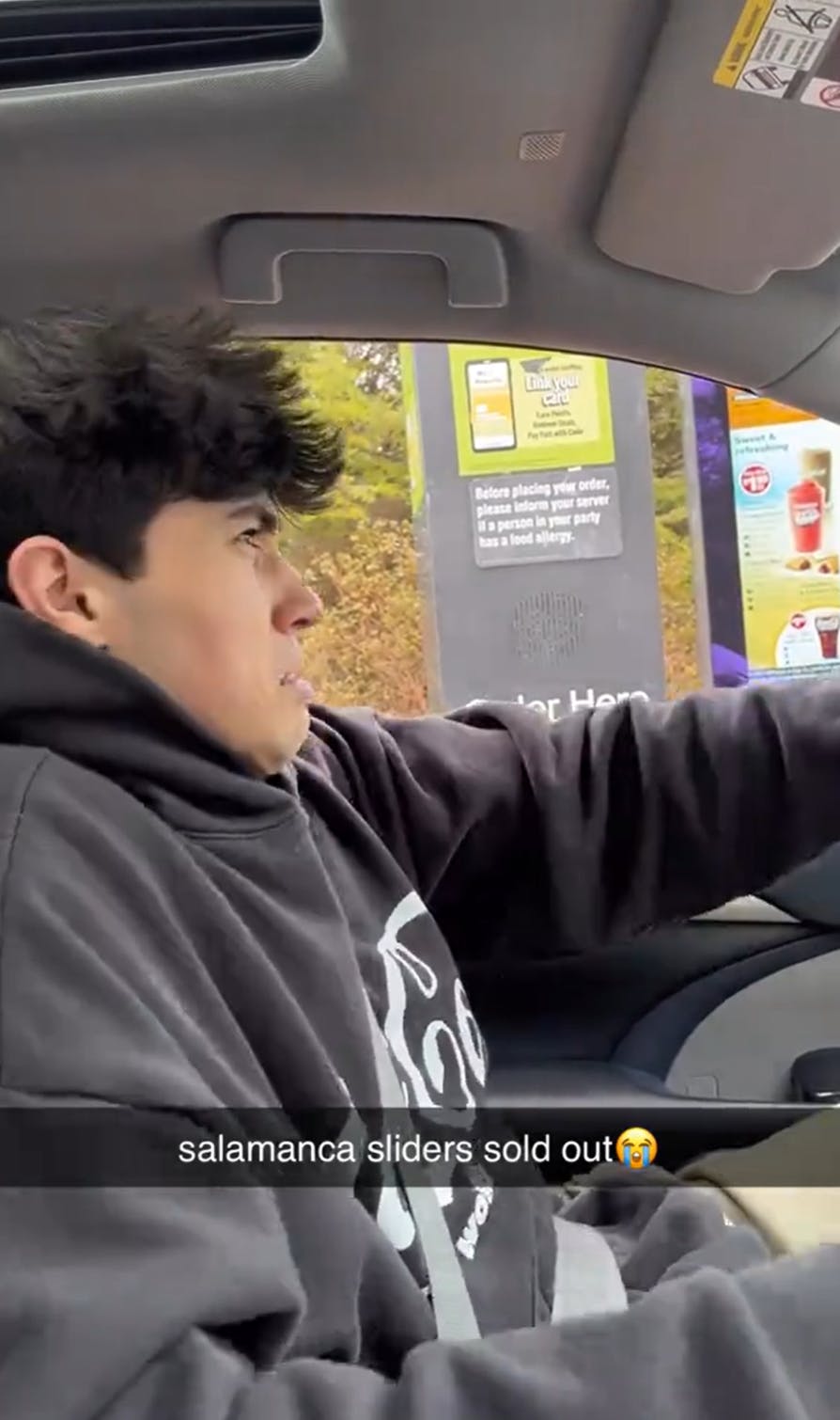 A young man sitting in the driver seat at a drive through looking disturbed. Text overlay reads, 'salamanca sliders sold out (sob emoji)'