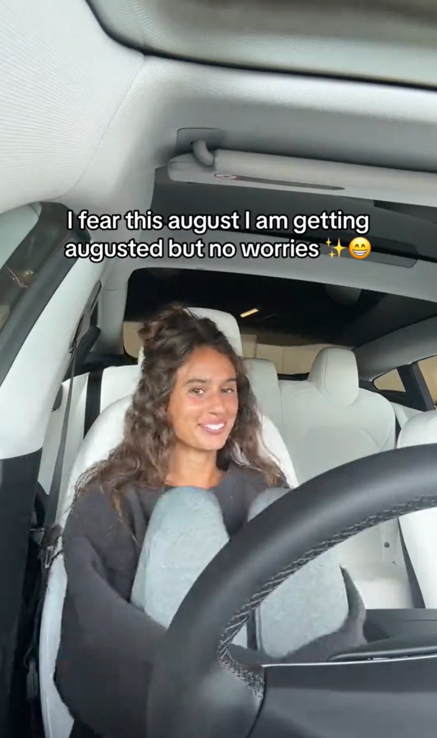 A woman sad but smiling for the camera sitting in her car with her knees up . Text overlay reads, 'I fear this august I am getting augusted but no worries'