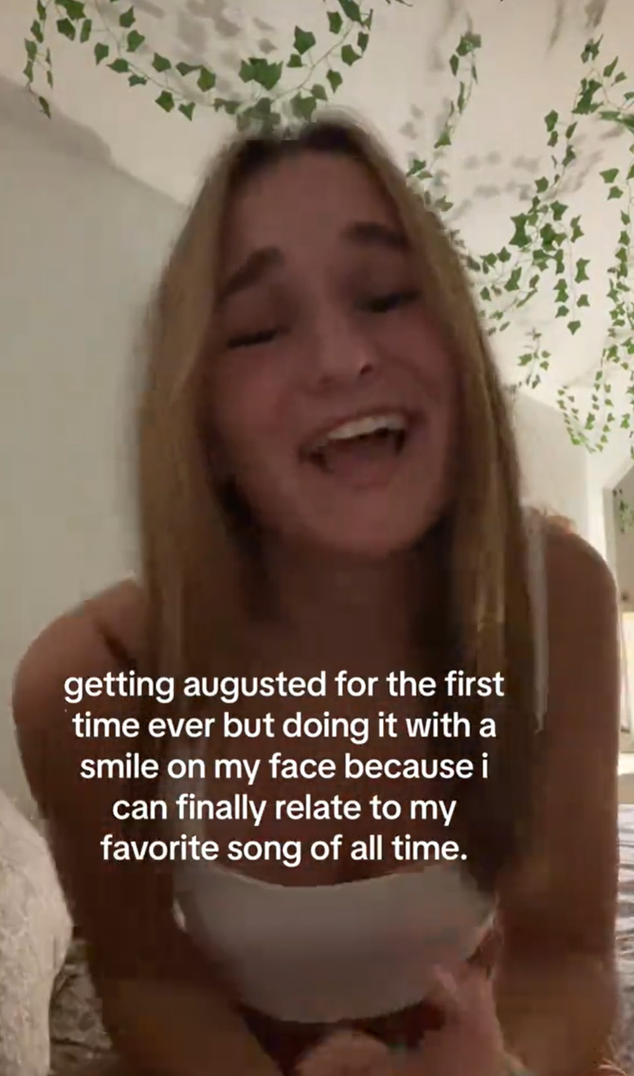 A blonde woman sitting in her bedroom singing for the camera and smiling. Text overlay reads, 'getting augusted for the first time ever but doing it with a smile on my face because i can finally relate to my favorite song of all time.'