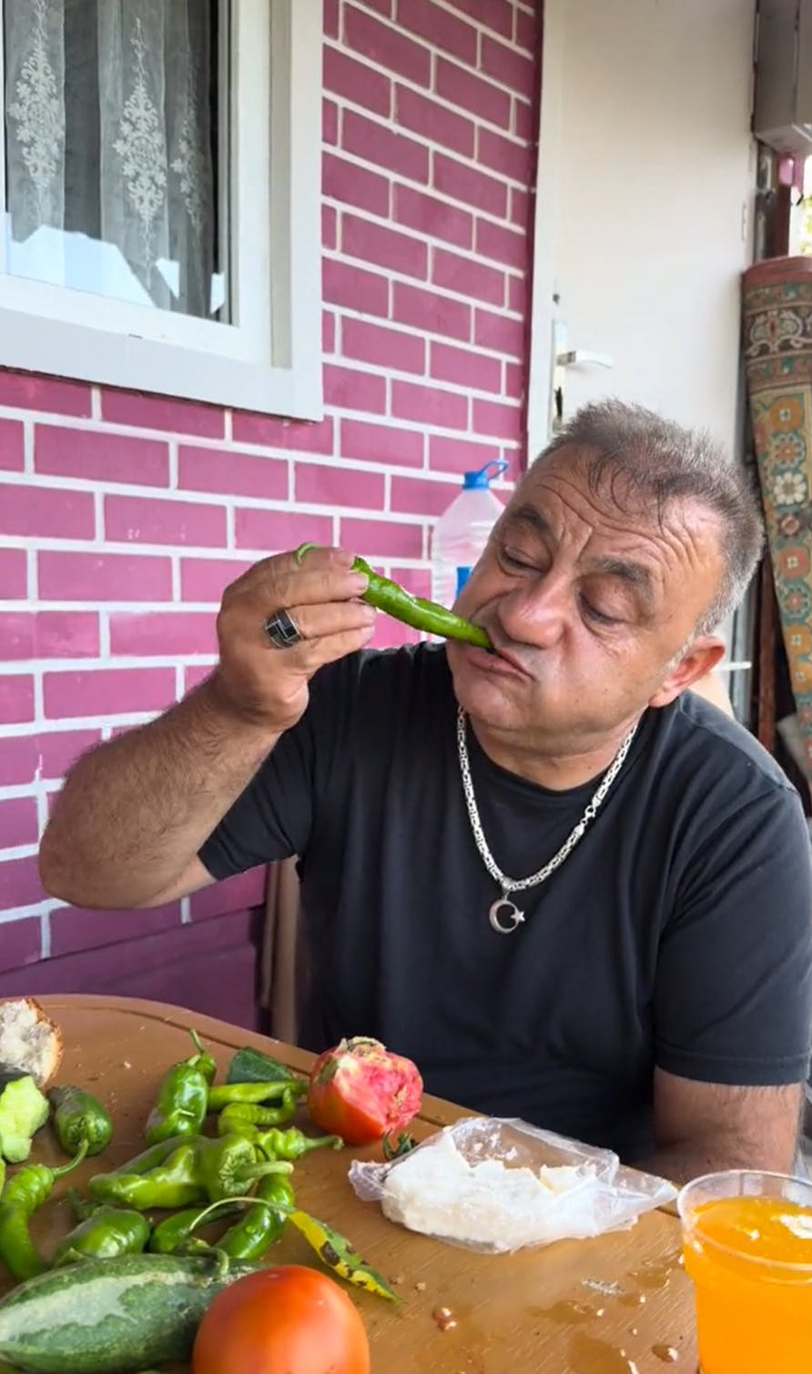 An older man sitting at a table outside, a pile of vegetables sitting in front of him for a Balkan Breakfast. He is biting into a raw pepper.