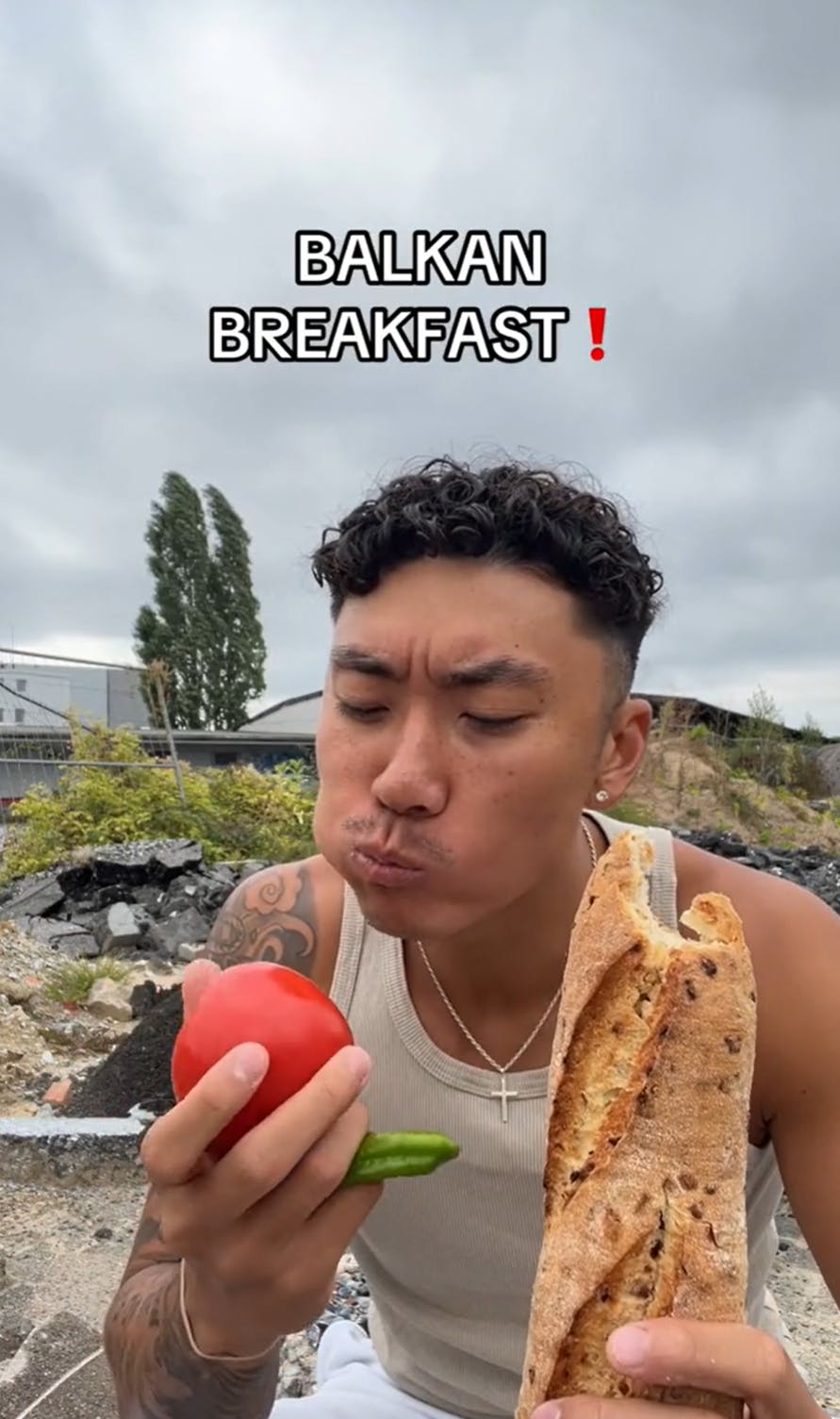 A man sitting on the ground outside, a baguette in one hand and a raw tomato and green pepper in the other. Text overlay reads, 'Balkan Breakfast!'