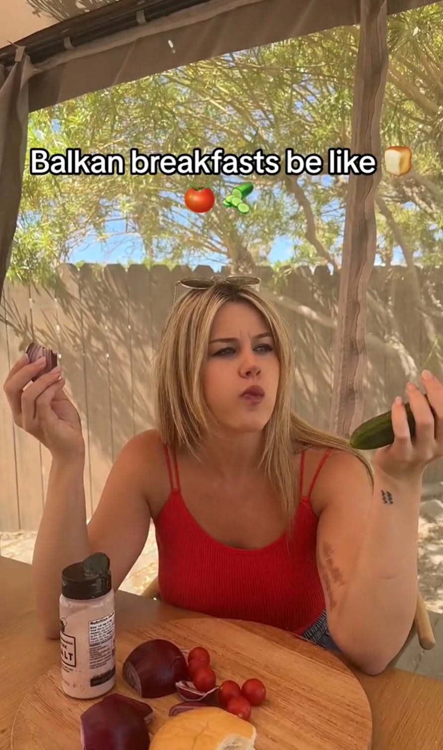 A woman sitting at a table outside staring off into the distance. She is holding a cucumber and just took a bite into a raw red onion. Text overlay reads, 'Balkan breakfasts be like (bread, tomato, cucumber emojis)'