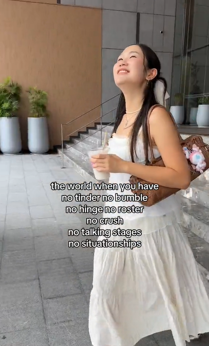 A woman in a white dress carrying a cold drink stares up at the sky with a wide grin on her face. Text overlay reads, 'the world when you have no tinder no bumble no hinge no roster no crush no talking stages no situationships.'